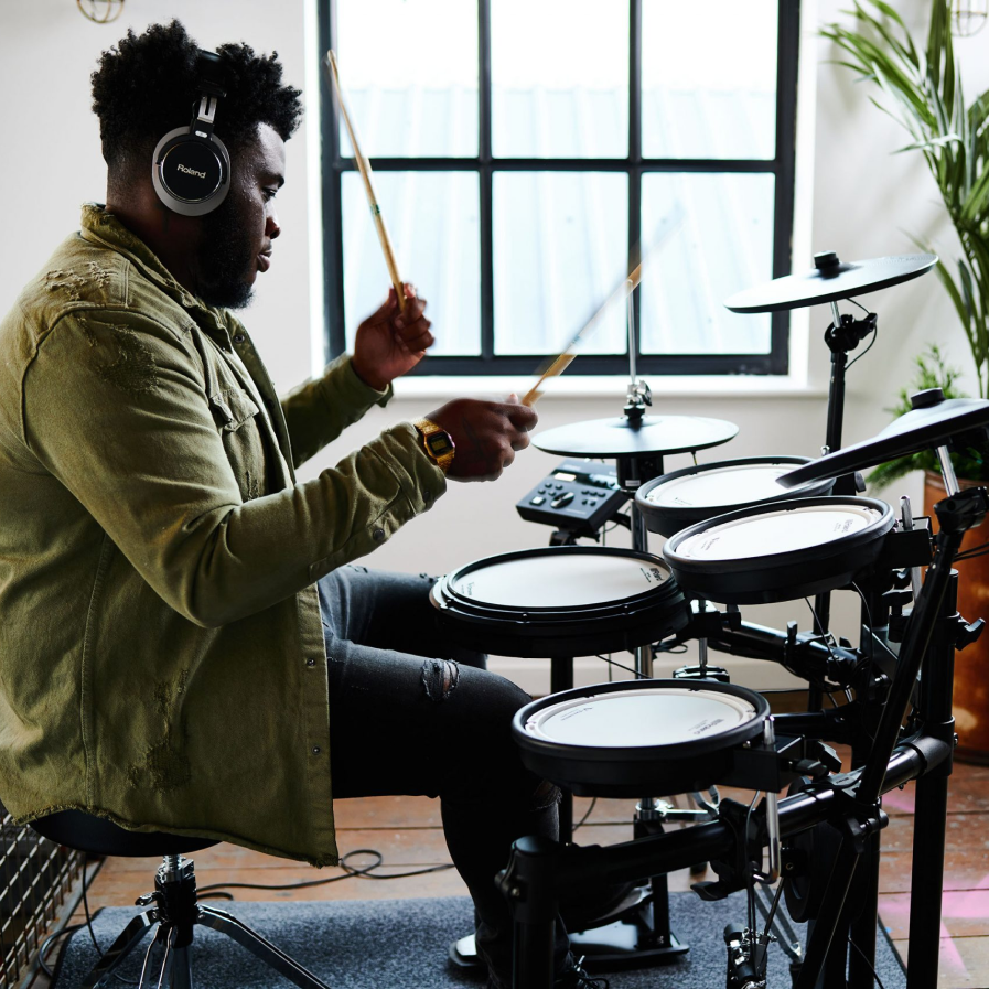 A man in a bright apartment playing a Roland electronic drum kit.