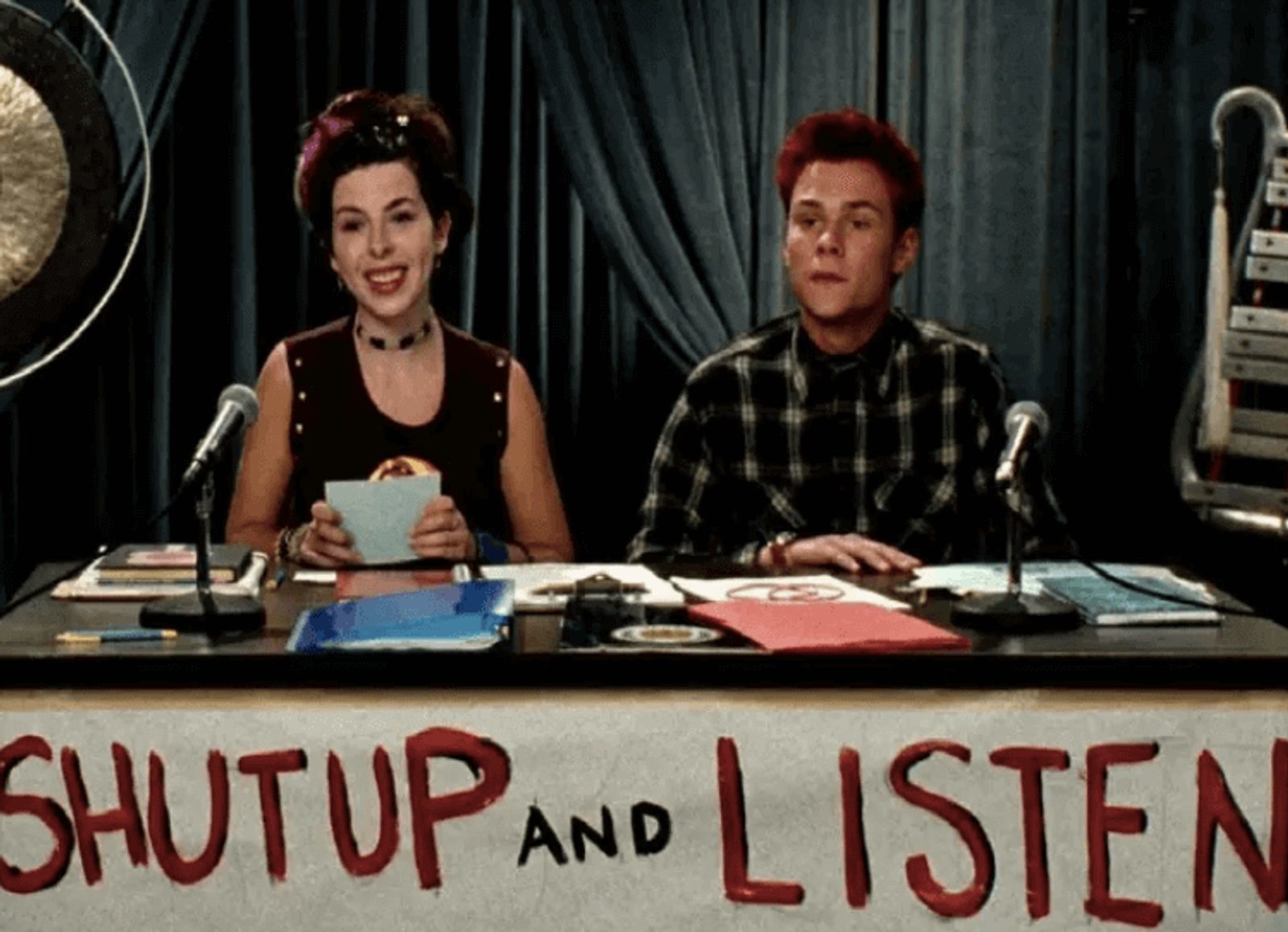 A man and woman sitting behind a table with a sign reading "shut up and listen"