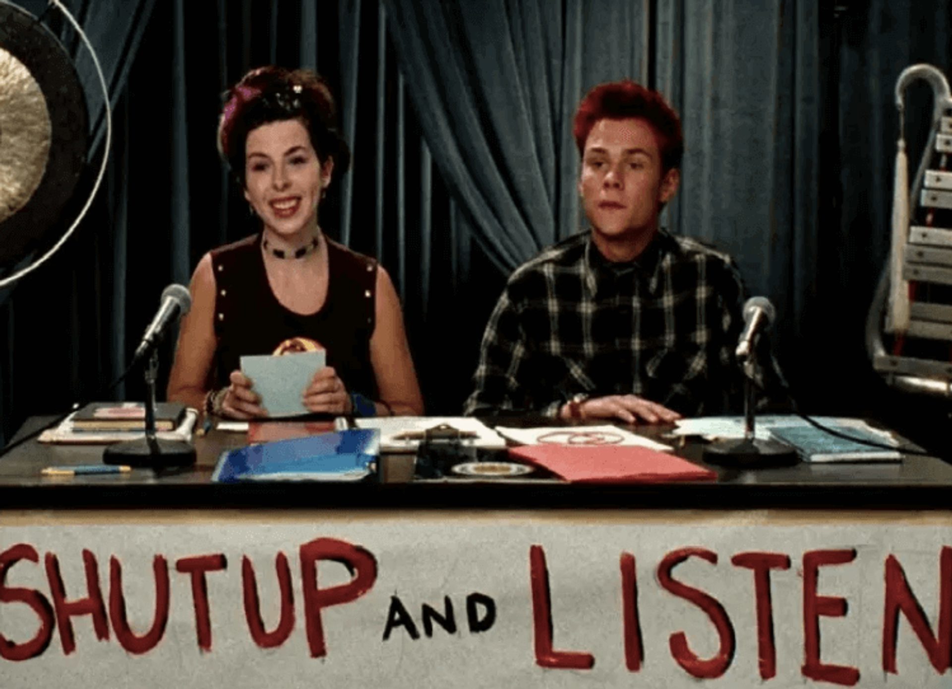 A man and woman sitting behind a table with a sign reading "shut up and listen"